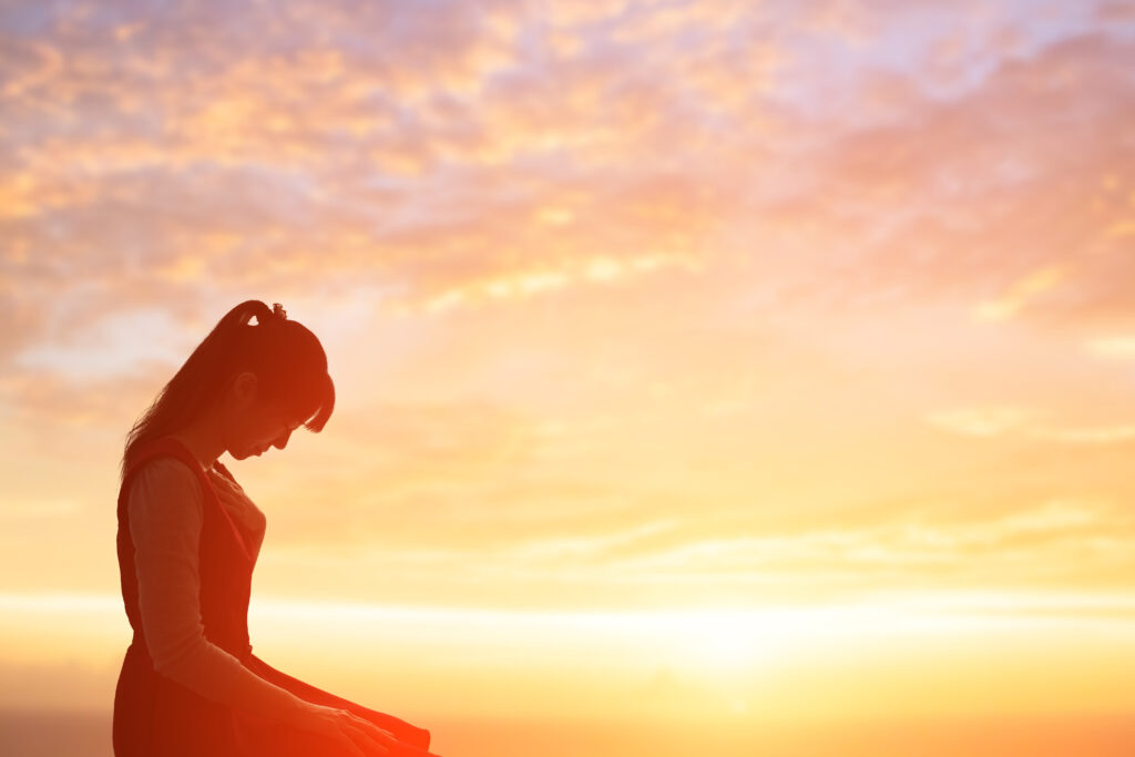 forgiveness pitfalls - A woman sitting on her knees looks down with her eyes closed as the sun sets behind her