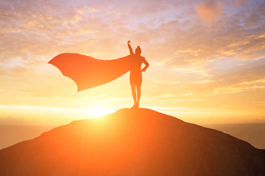 A person wearing a cape standing on top of a hill with their fist raised up, starting their hero’s journey