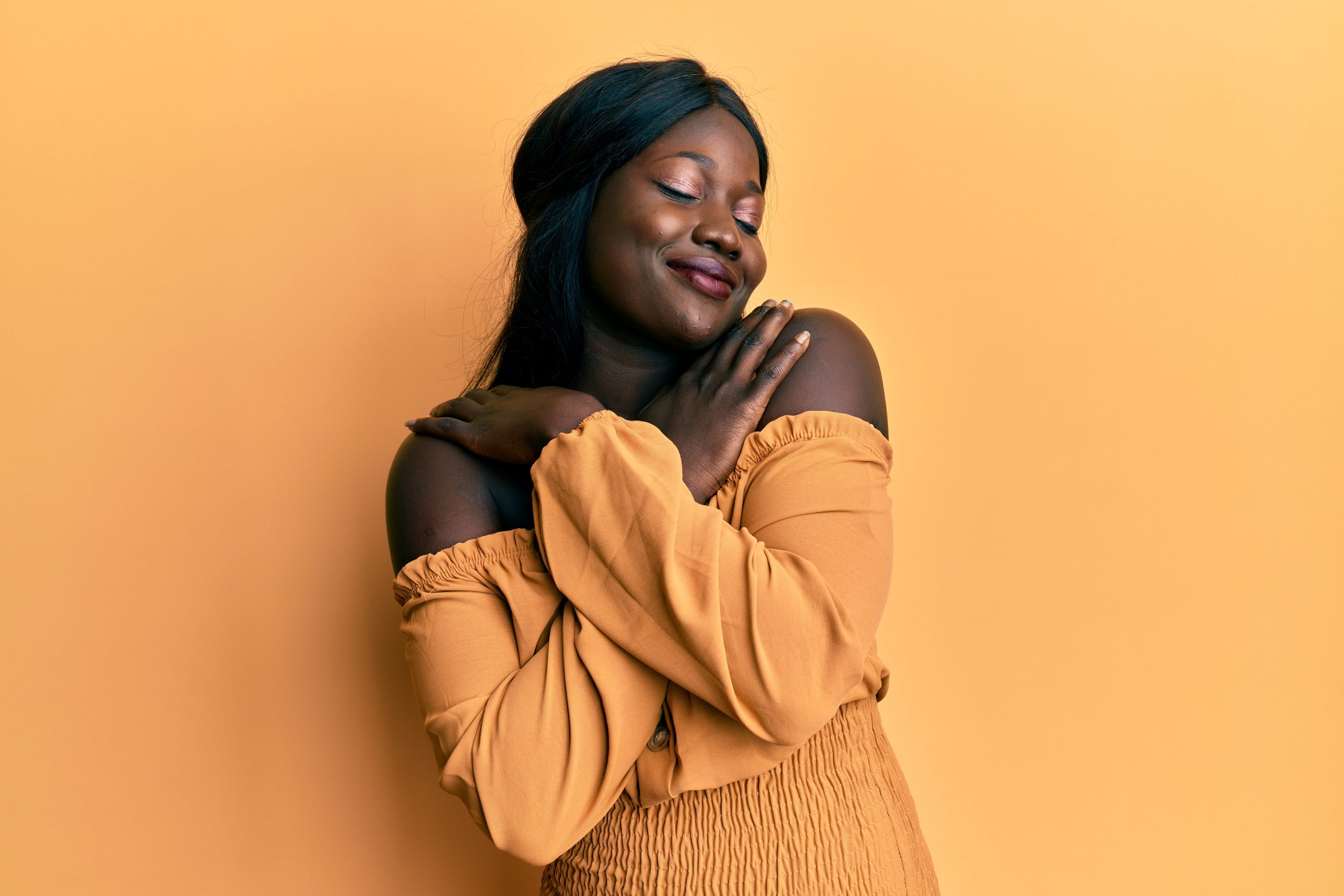 A woman in a yellow dress smiling and hugging herself, experiencing self love